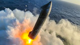 a black rocket with wing-like canards fires its engines just above the ocean, creating a spray of water vapor