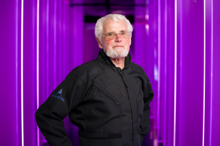 An older man with white hair and beard stands for a portrait photo wearing a black coat against a purple background.