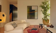 Beige living room with curved cream sofa, soft red-brown rug, red round coffee table and large plant