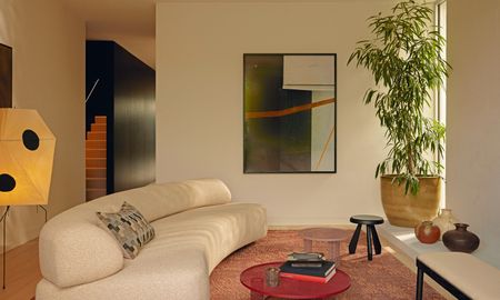 Beige living room with curved cream sofa, soft red-brown rug, red round coffee table and large plant