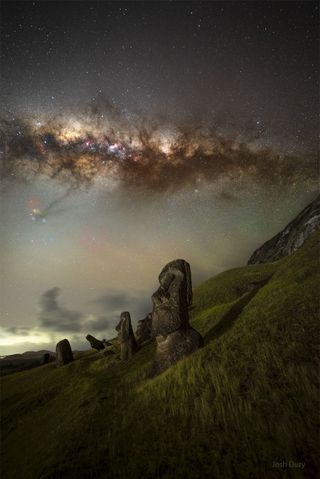 Large statues of giant heads stand on a grassy hillside beneath a night sky illuminated with brilliant yellows and oranges from the Milky Way, masked by the cosmic shadow of space among the stars.