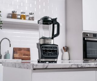 A Ninja blender on a marble countertop; in the background, a sink, shelves, and an oven against white walls.