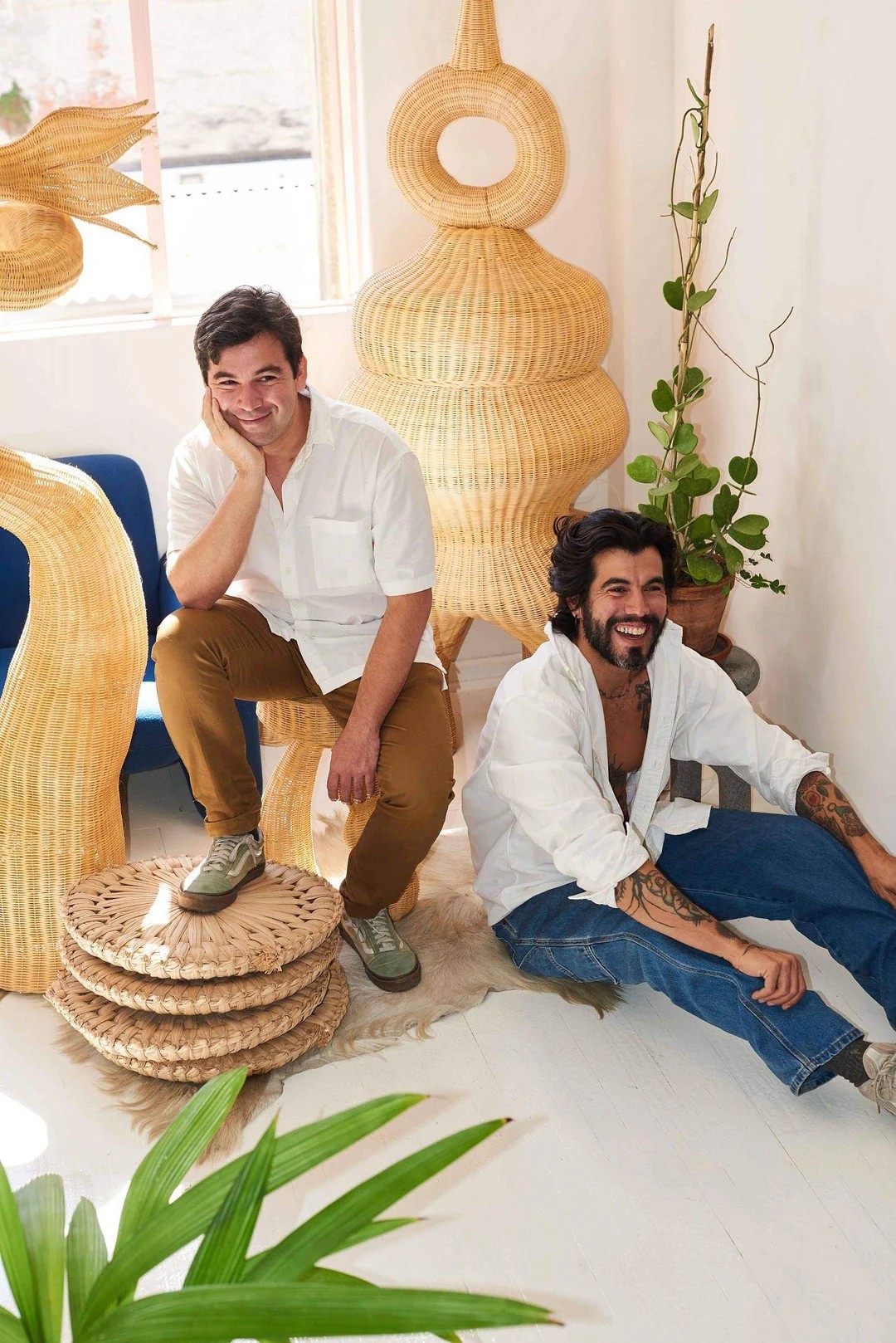 Two young men with white shirts and brown and denim trousers, respectively, smile at the camera in a sun-lit studio with white-washed wooden floors and lots of sculptural rattan creations.