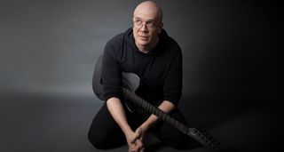 A colour portrait of Devin Townsend, crouched with his Aristides black T-Style electric guitar