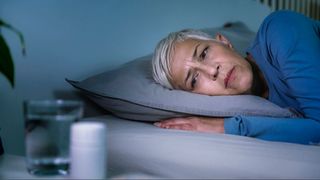 A woman with grey hair and a blue top laying on a pillow looking as glass of water and supplements on bedside table as she struggles with sleep during menopause