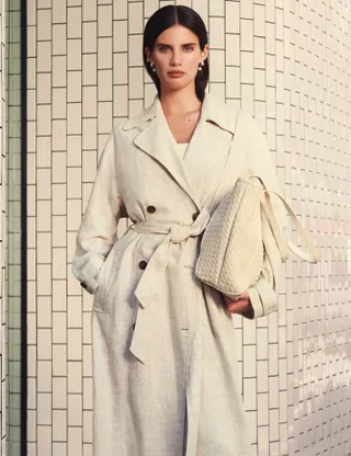 Model wears a cream linen trench coat, holds bag 