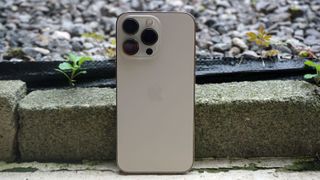 En bild på en vit iPhone 13 Pro som står lutad mot en tegelstenskant vid en rabatt, med baksidan mot kameran.