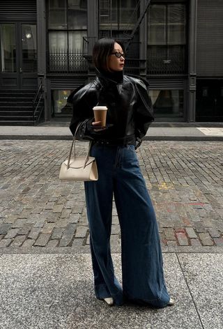 A woman wearing a black leather moto jacket with wide leg jeans, white pointed ankle boots, and a white shoulder bag.