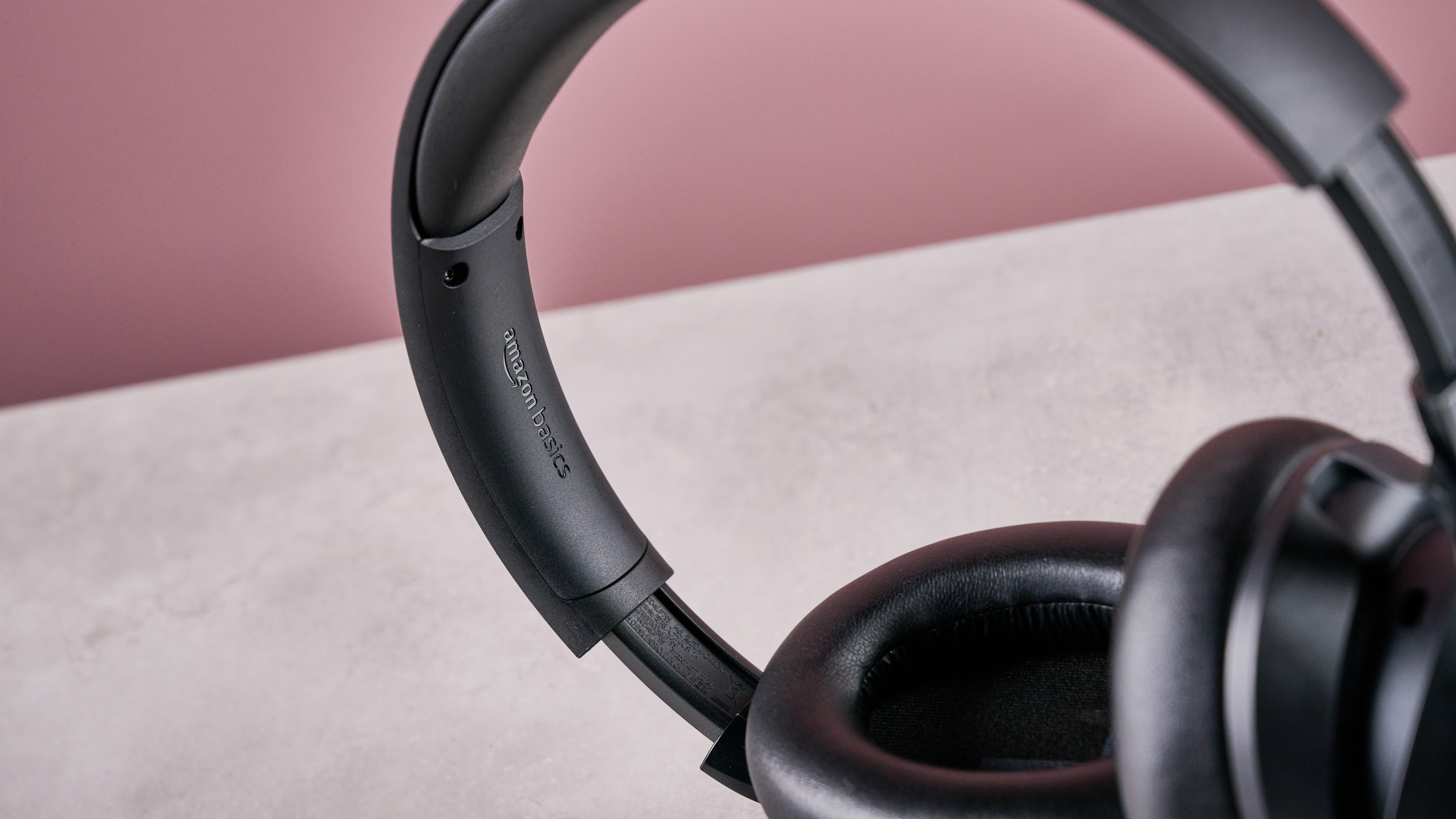 Amazon Basics Hybrid Active Noise Cancelling Headphones on a gray surface, with a pink background, showing the logo on the headband