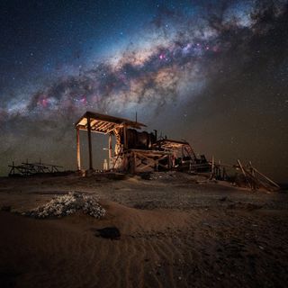 An abandoned mining structure in the desert stands, dimly illuminated under the streak of the Milky way, centered in the starry night sky.
