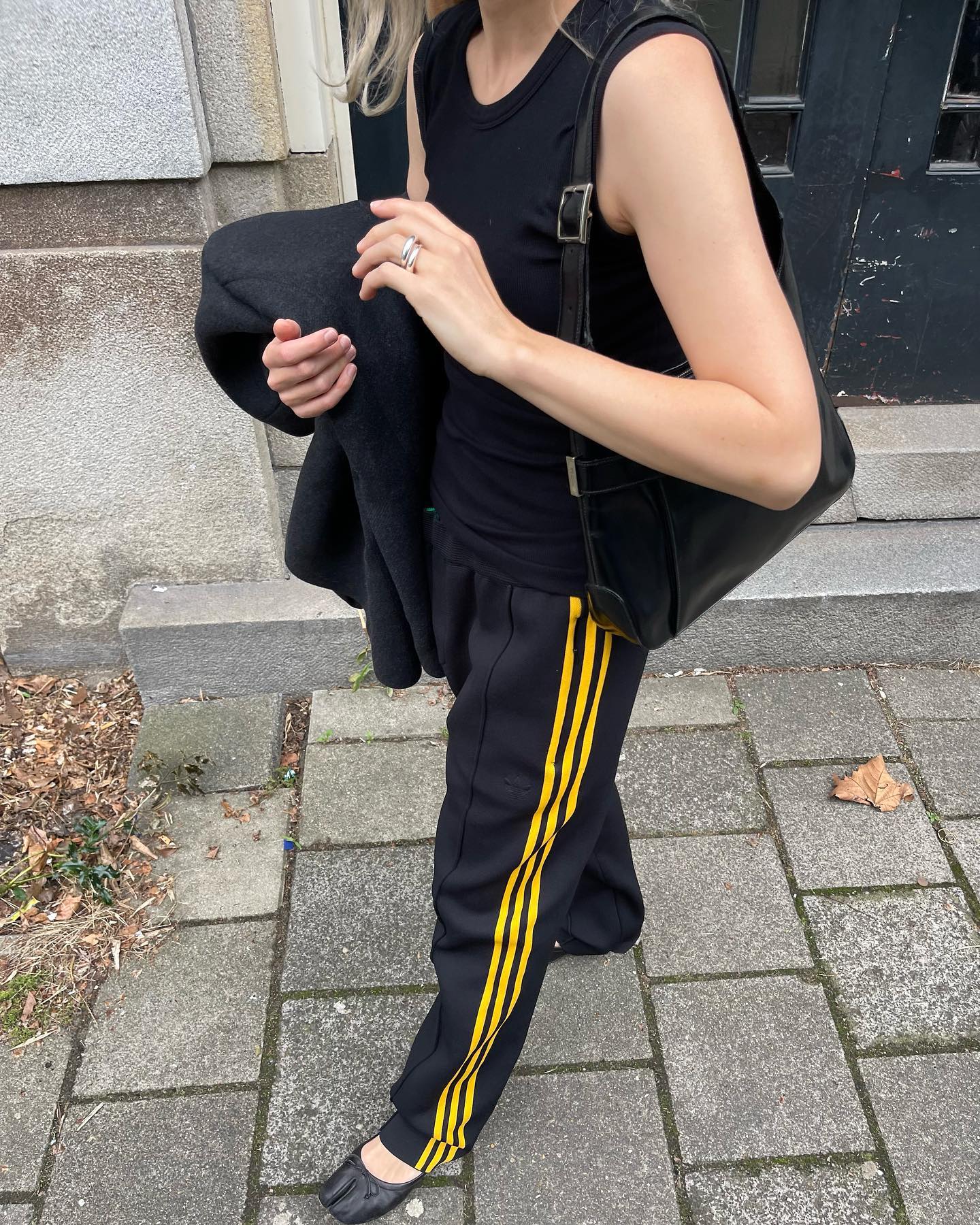Dutch style influencer Stephanie Broek walking on the sidewalk wearing a black tank top, black Adidas track pants with yellow stripes, and black Margiela ballet flats