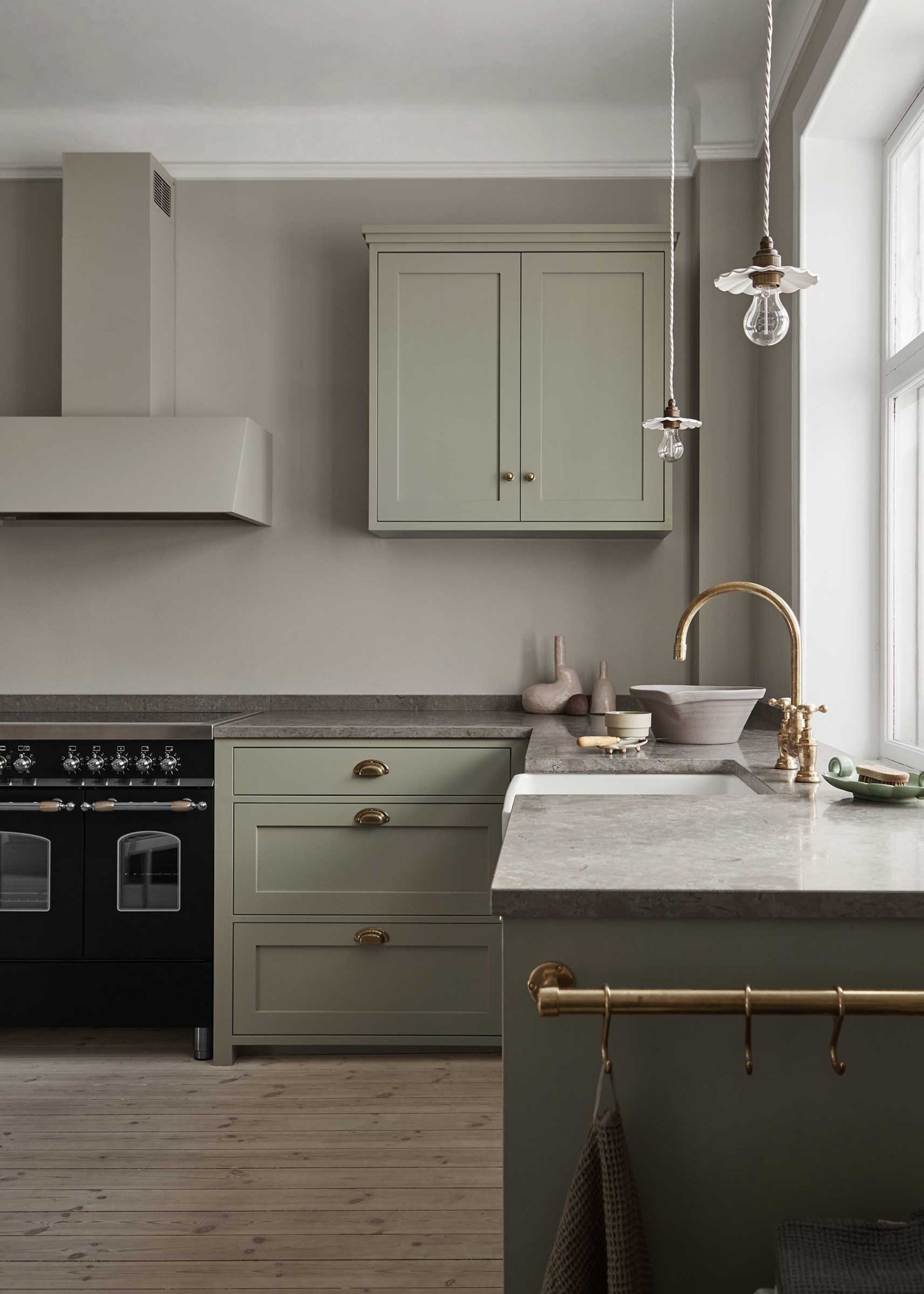 A pale green kitchen with a gas oven on the far left in black as well as a view of the kitchen sink and golden tap