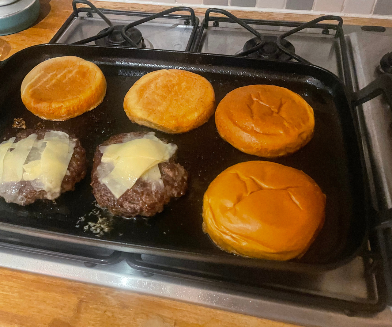 Two burger patties with melted cheese, and four buns, frying on a flat cast iron griddle.