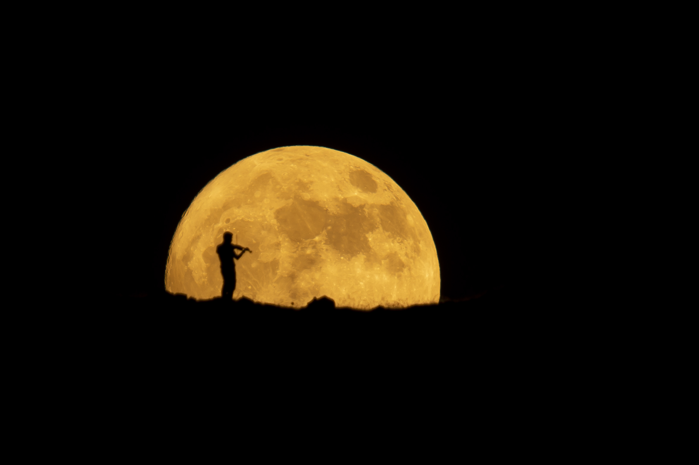 A photograph of the Strawberry Moon rising over the horizon in Iraq while a man plays the violin.