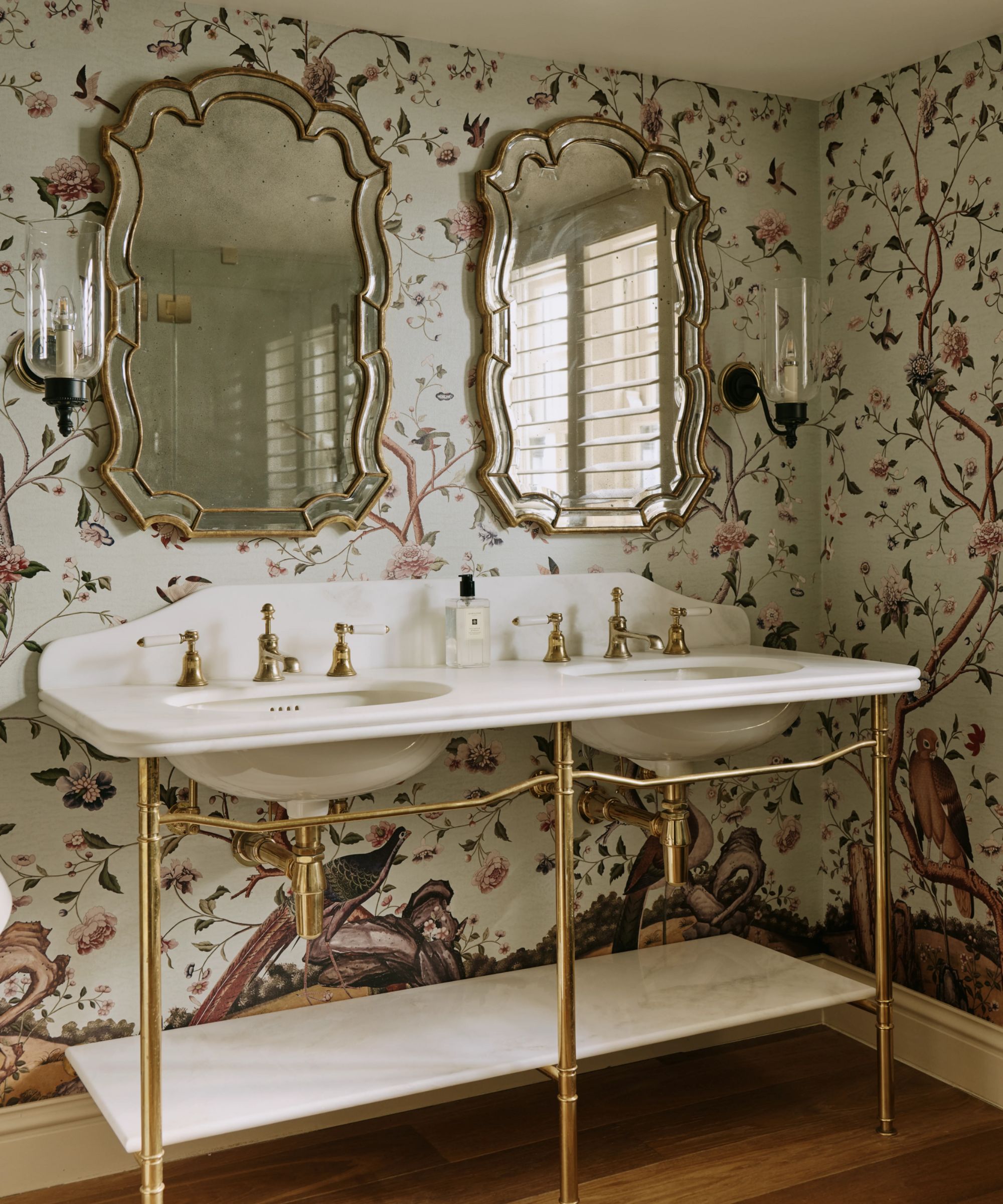 a vintage style bathroom with floral mural wallpaper, a victorian style double vanity with brass legs, and two antique mirrors