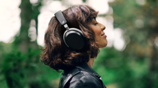 A woman in a black jacket wearing Sennheiser HDB 630 headphones with trees and bushes blurred in the background