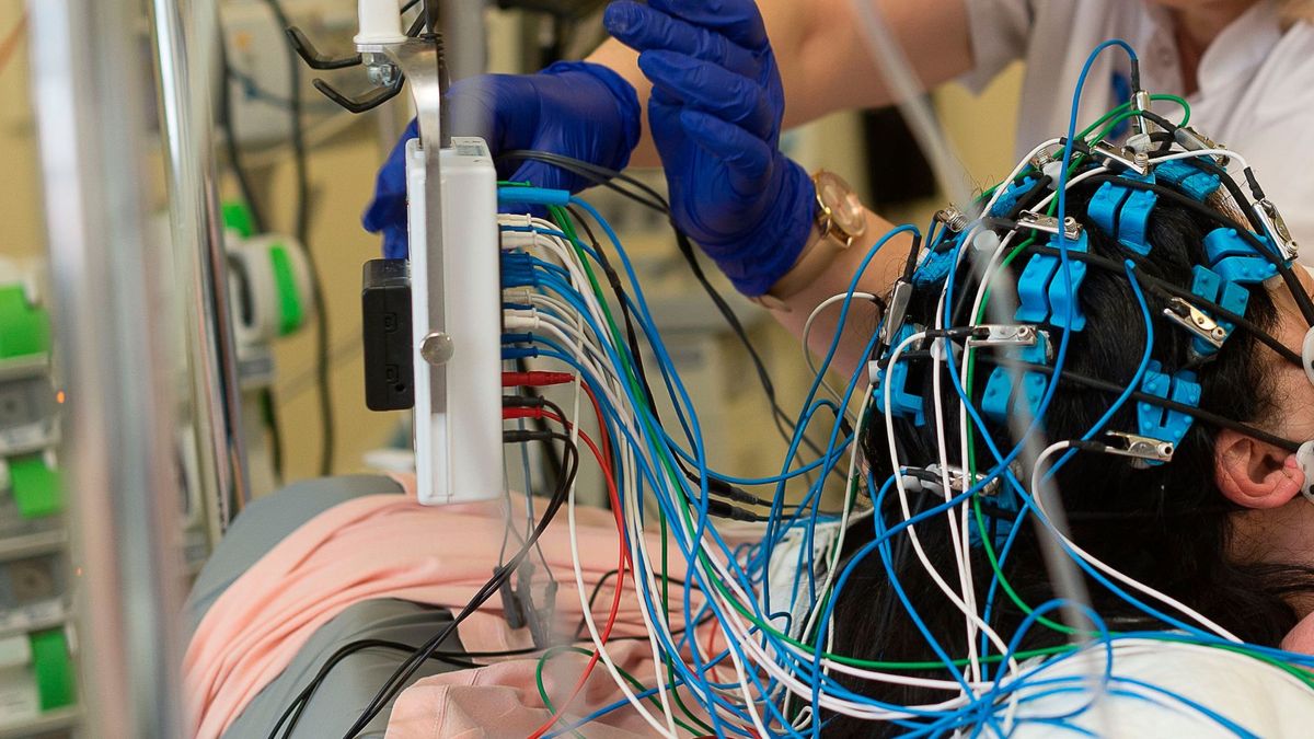 close up photo of a medical provider&#039;s gloved hands as they adjust colorful wires that are stuck to a hospitalized patient&#039;s head