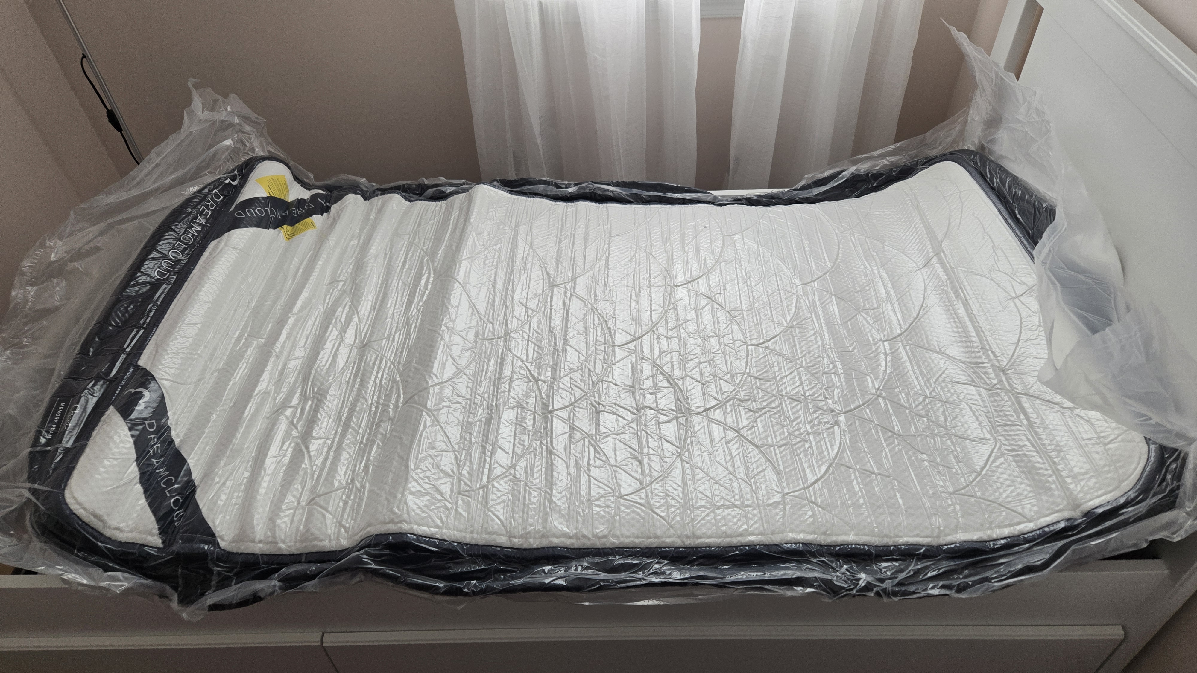DreamCloud Classic Memory Foam Mattress unrolled but still flat and wrapped in plastic on a platform bed