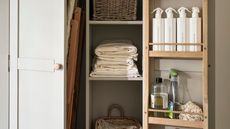 An open closet with door mounted storage with cleaning spray bottles inside. Folded linens on a shelf between tow shelves with baskets on them. An ironing board leaning against a middle divider partially hidden behind a second door. 