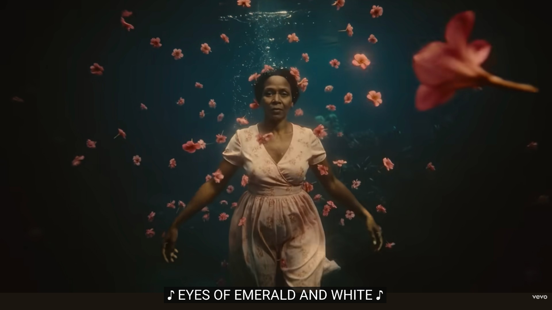 A woman with dark skin and short hair wears a pale pink dress and is submerged underwater, floating amidst numerous pink and orange flowers. The background is dark, and the image includes a caption overlay on the bottom that reads: &quot;&amp;#127925; EYES OF EMERALD AND WHITE&quot;.