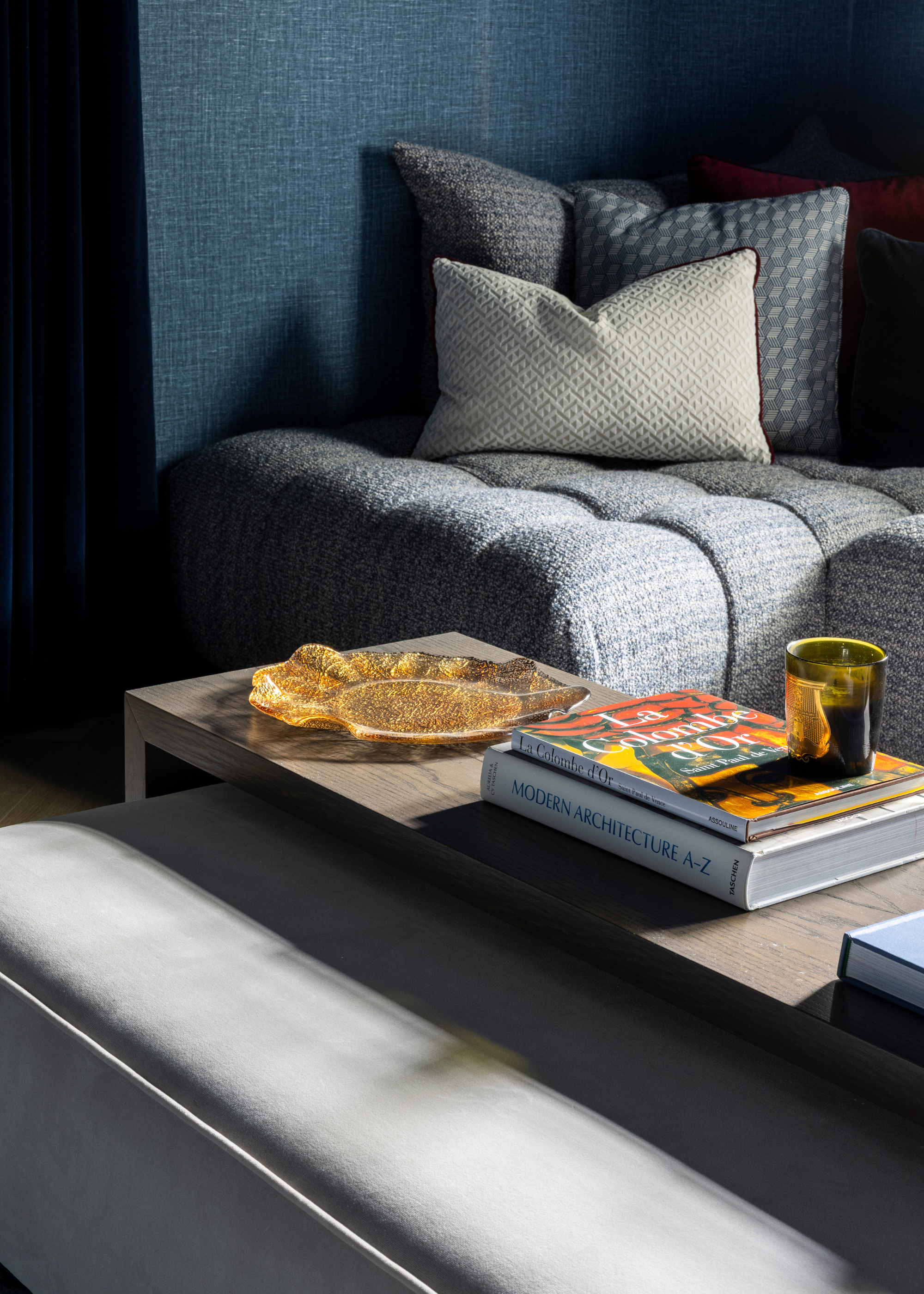 A coffee table over the center of a gray ottoman styled with books, a gold decorative tray and a candle with a gray sofa in the background against a dark blue wall styled with cushions