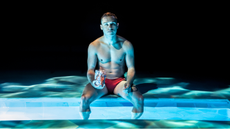Finn Cole as Ray sitting by the pool in Red Speedo at the Orange Tree Threatre.