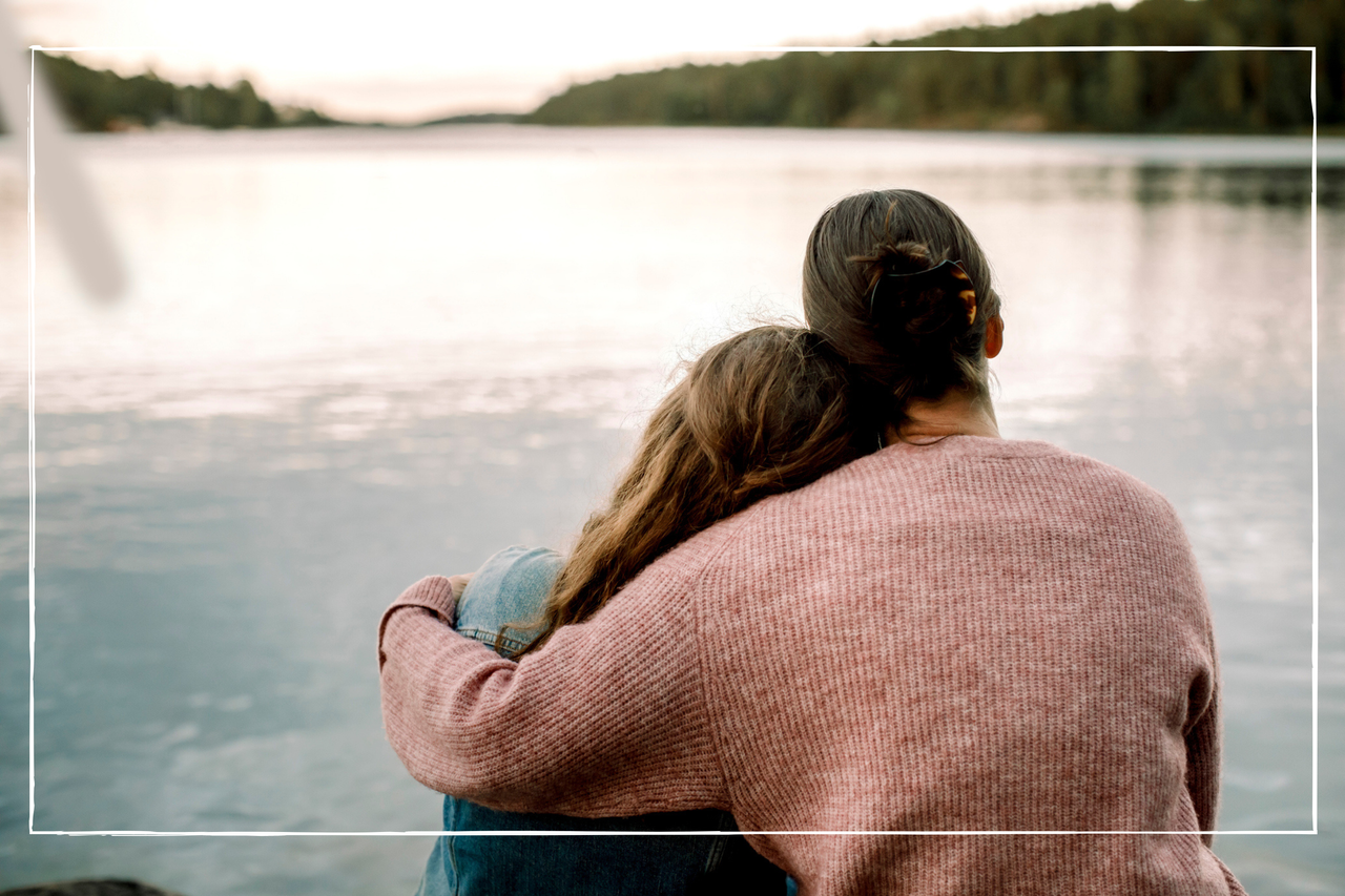 A mother with her arm around her daughter, who is leaning on her shoulder - they&#039;re looking out onto a pond 