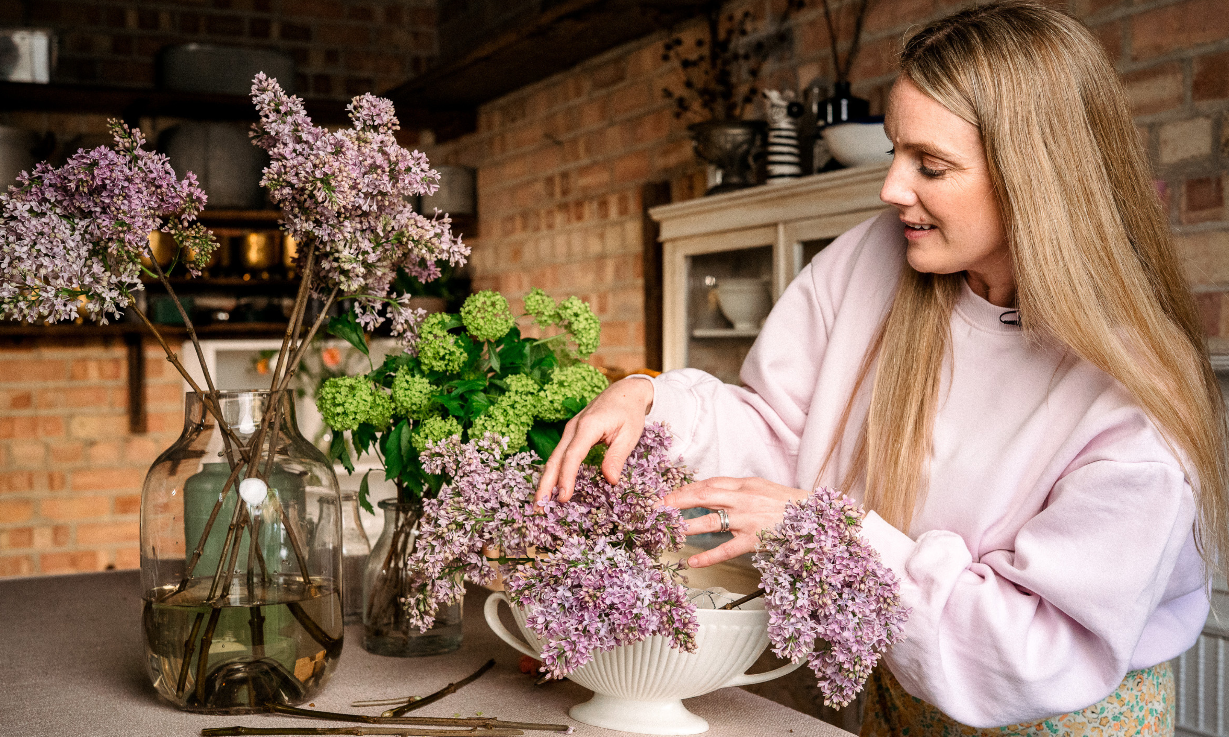 woman in pale pink jumper placing lilac stems into white boat vase making a floral table design