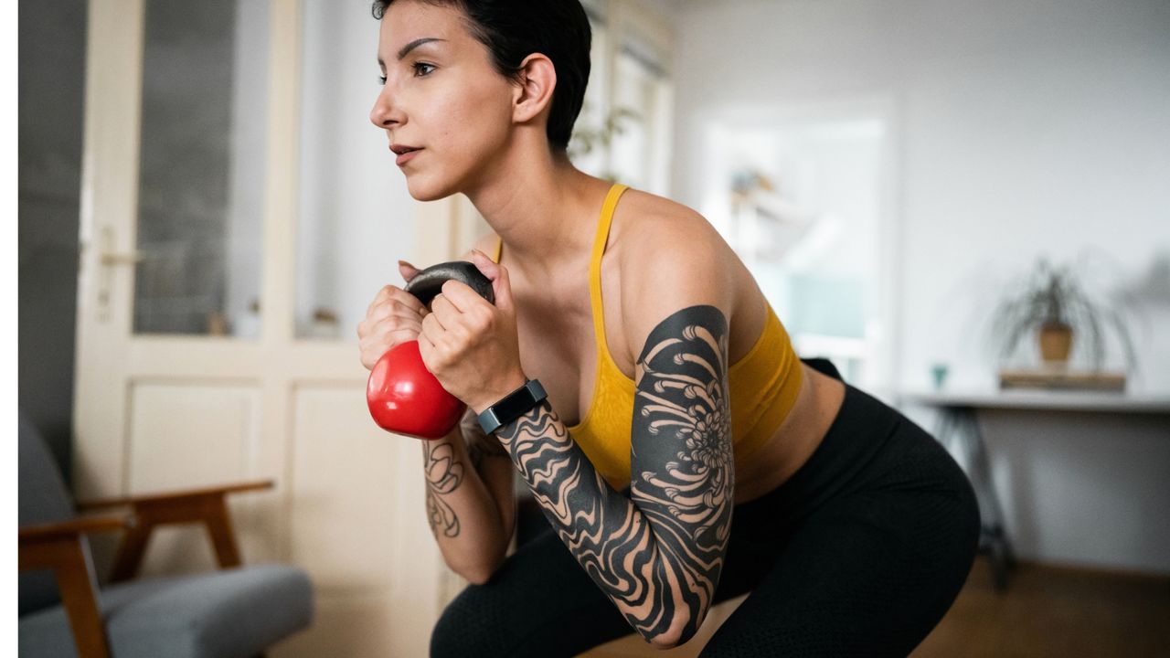 woman wearing a yellow vest and black trousers with tattoos down one arm doing a kettlebell squat with a small red kettlebell in a home setting. 