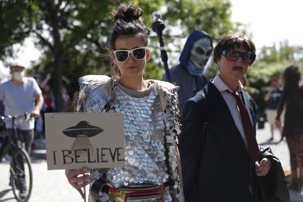 A woman holds a banner with an UFO painted on it and reading &amp;quot;I believe&amp;quot; as another protester dressed up as alien walks by at a demonstration against conspiracy theorists at Mauerpark, amidst