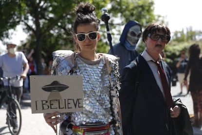 A woman holds a banner with an UFO painted on it and reading "I believe" as another protester dressed up as alien walks by at a demonstration against conspiracy theorists at Mauerpark, amidst