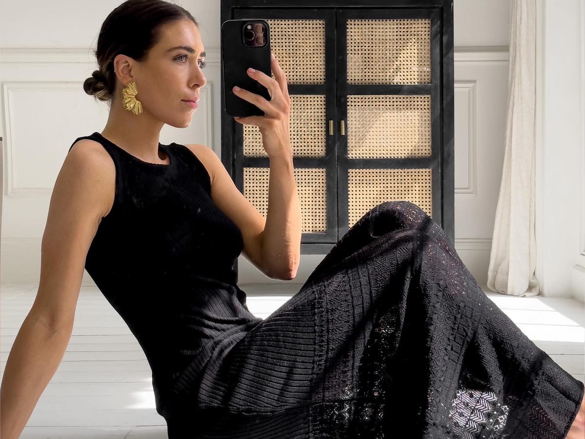 stylish woman sits on white wood floor posing for a mirror selfie wearing statement gold earrings and a black embroidered dress