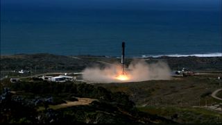 a scorched rocket lands on a launch pad surrounded by green foliage and the sea