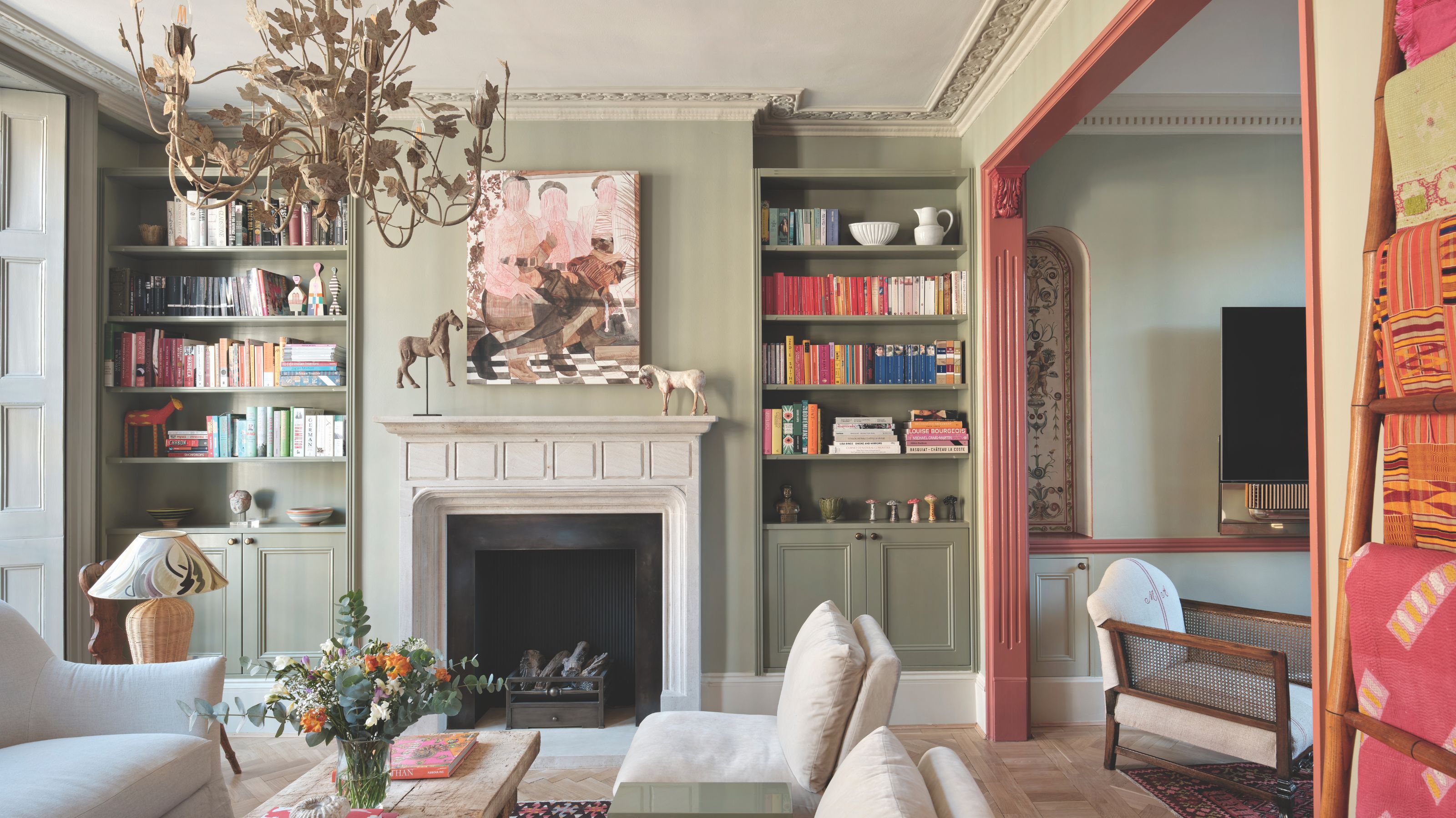 A sophisticated, eclectic living room features a stone fireplace, built-in green bookshelves filled with books, a large abstract painting, and a grand, aged gold chandelier. A terracotta-painted archway leads to an adjacent room, next to a ladder displaying colorful textiles.