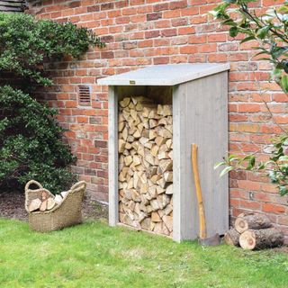 firewood store with logs outside next to a wall, basket of logs next to it, lawn, axe