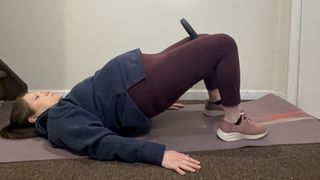 Jennifer Rizzuto performs a glute bridge with a Pilates ring in her home on a mat. She is on her back, with her feet planted on the floor, knees bent and hips elevated off the ground. The Pilates ring is being squeezed between her thighs.