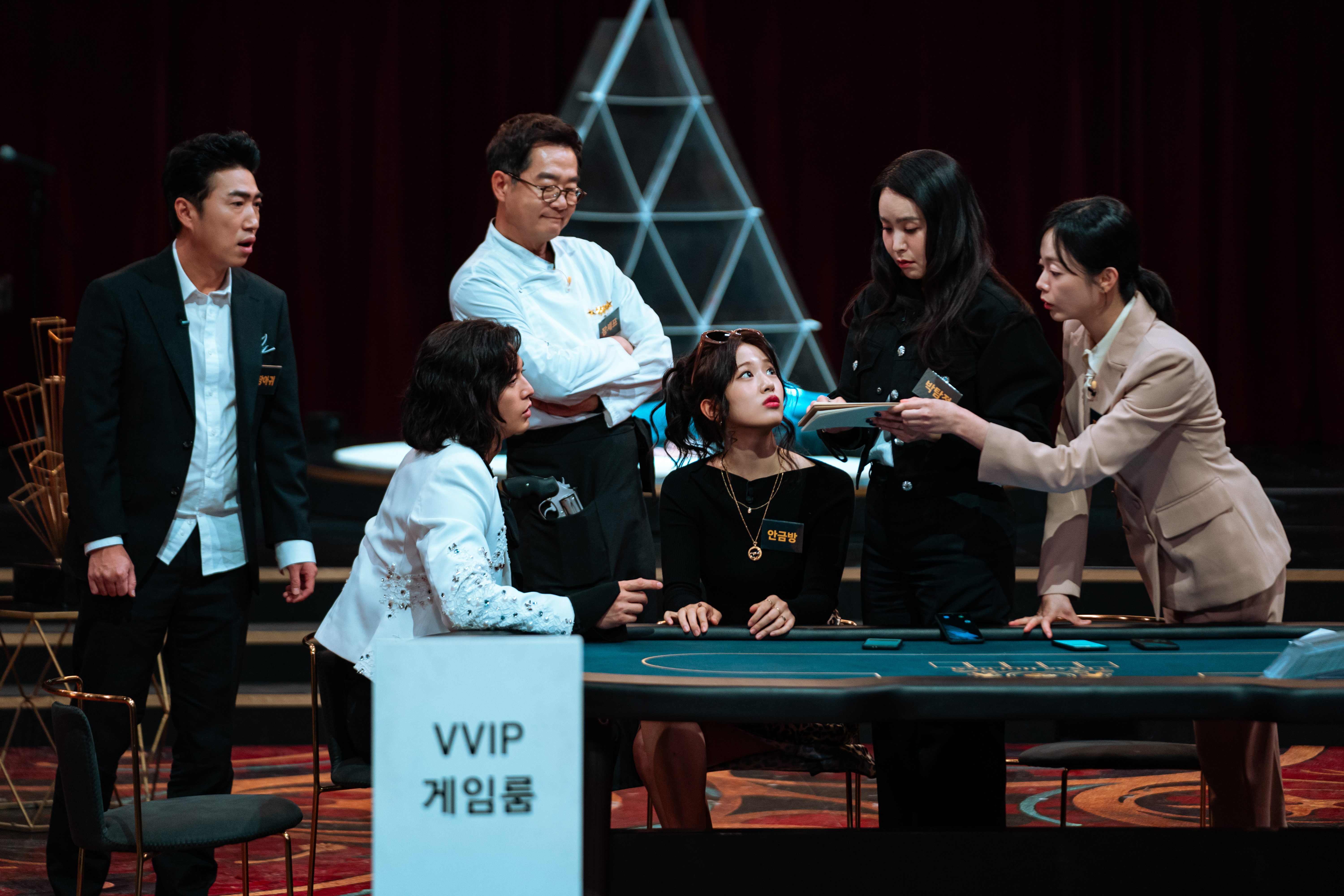 A group of people (l-r Jang Dong-min, Kim Ji-hun, Jang Jin, An Yu-jin, Park Ji-yoon, Jeon So-min) discuss the contents of a notebook while standing and sitting around a VVIP poker table, in Netflix&#039;s &#039;Crime Scene Zero.&#039;