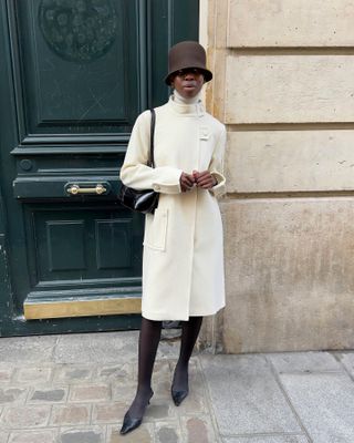 Sylvie Mus wearing a cream coat with tights, black heels, a black shoulder bag, and a brown cloche hat.