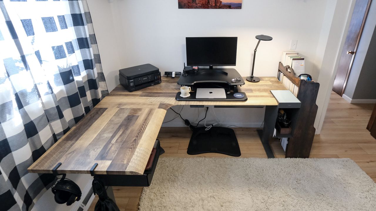 The Vari L-Shape Electric Standing Desk ina corner of a home office with a laptop, monitor, printer and other work from home accessories