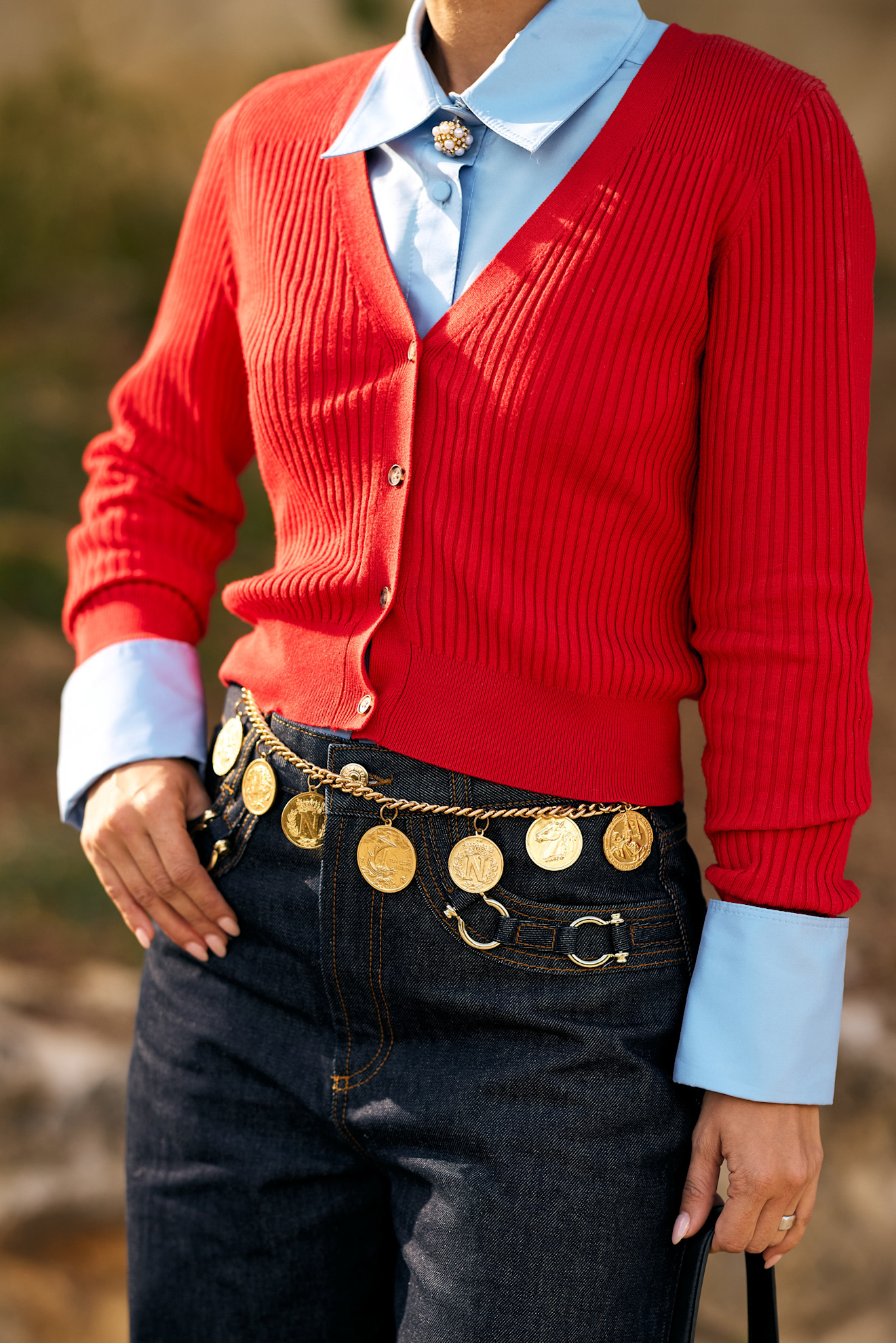 a white woman close up to her torso with a red sweater, blue shirt, gold belt and jeans