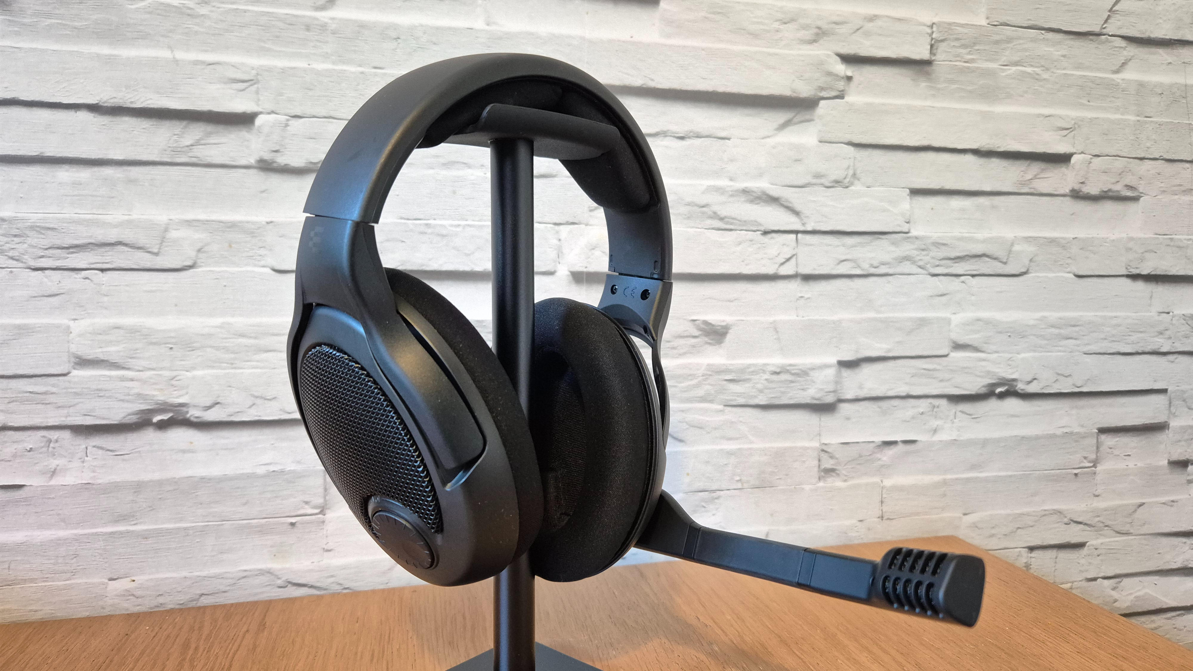 The Drop + EPOS PC38X headset on a headset stand with a white brick background and wooden surface underneath