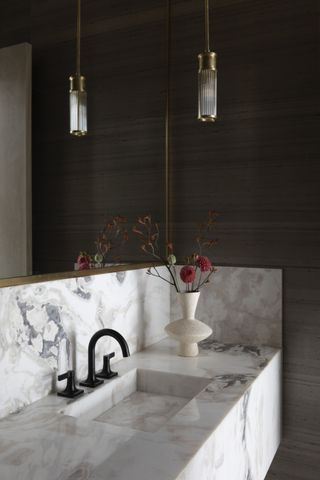 A neutral bathroom with a marble vanity and sink, dark brass taps, black panelled wall, hanging glass pendant and a vase with flowers