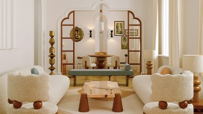 A white living room with textured sofa chairs, long white sofas, a low coffee table, a blue bench and a craved doorway leading to a dining room with a wall of framed paintings and wall sconces