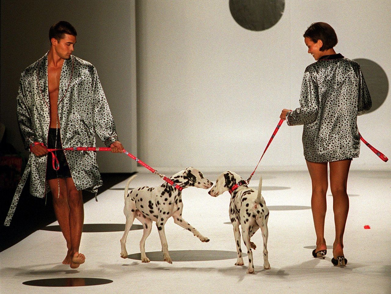 Two fashion models walk dalmatians on leads down a catwalk, wearing spotted garments that echo the dogs&rsquo; coats.