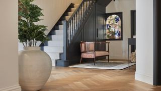 entrance hall with wooden flooring, grey staircase with cream runner, wooden chair on rug, stained glass window and large cream plant pot