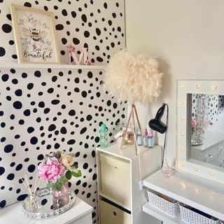 a girly room with dalmation polka dot wallpaper, a white dressing table with a mirror above that has a white frame and lights, storage cubes in the corner and a dresser against the wall which has a vase and incense sitting on top