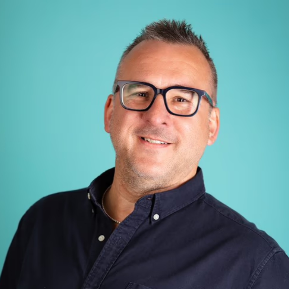 A man wearing thick rimmed glasses and a navy shirt smiles at the camera against a teal backdrop