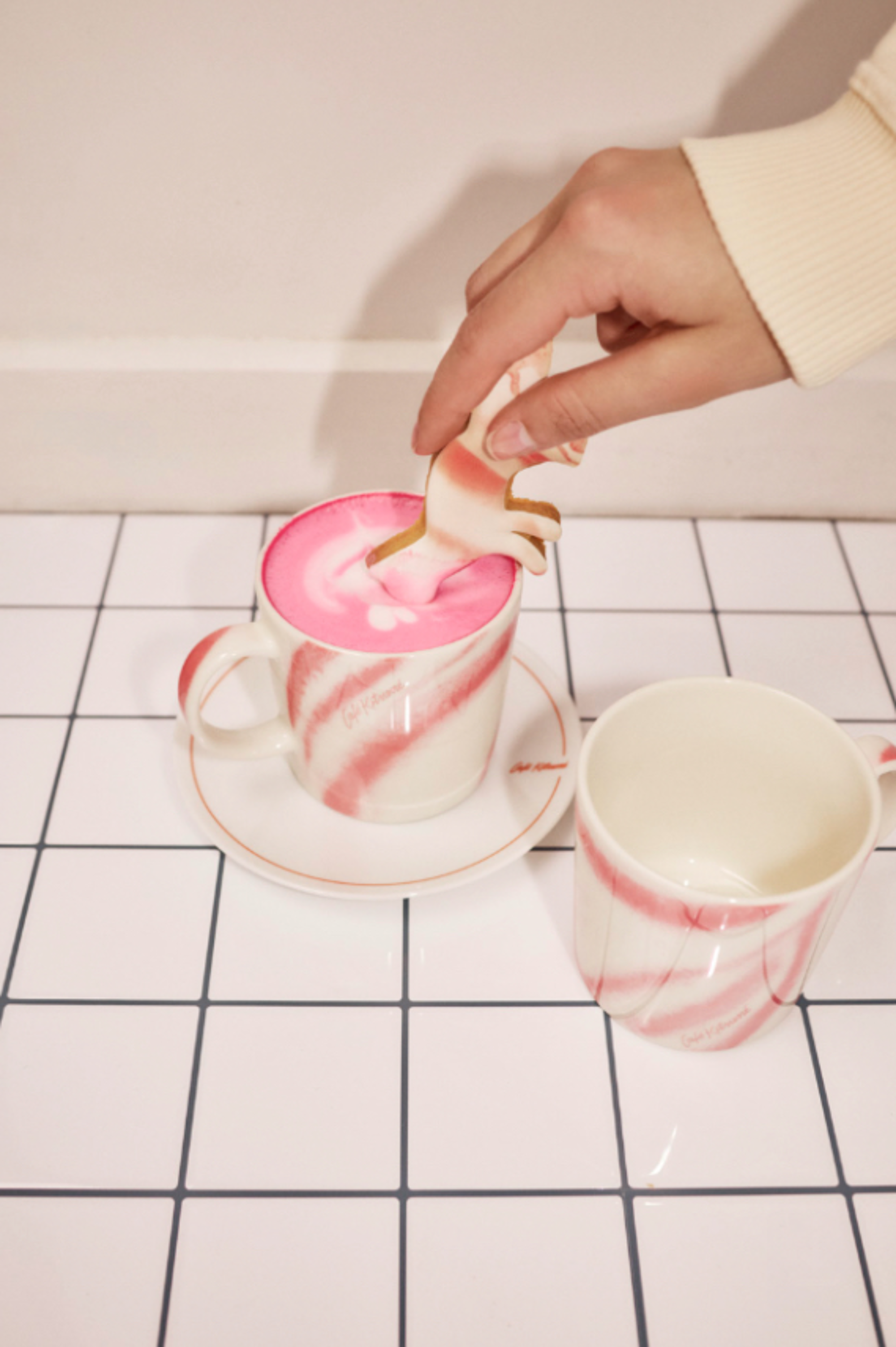Image of a hand holding a cow cookie and dunking it into a pink coffee inside a pink and white swirl mug on a white tile countertop.