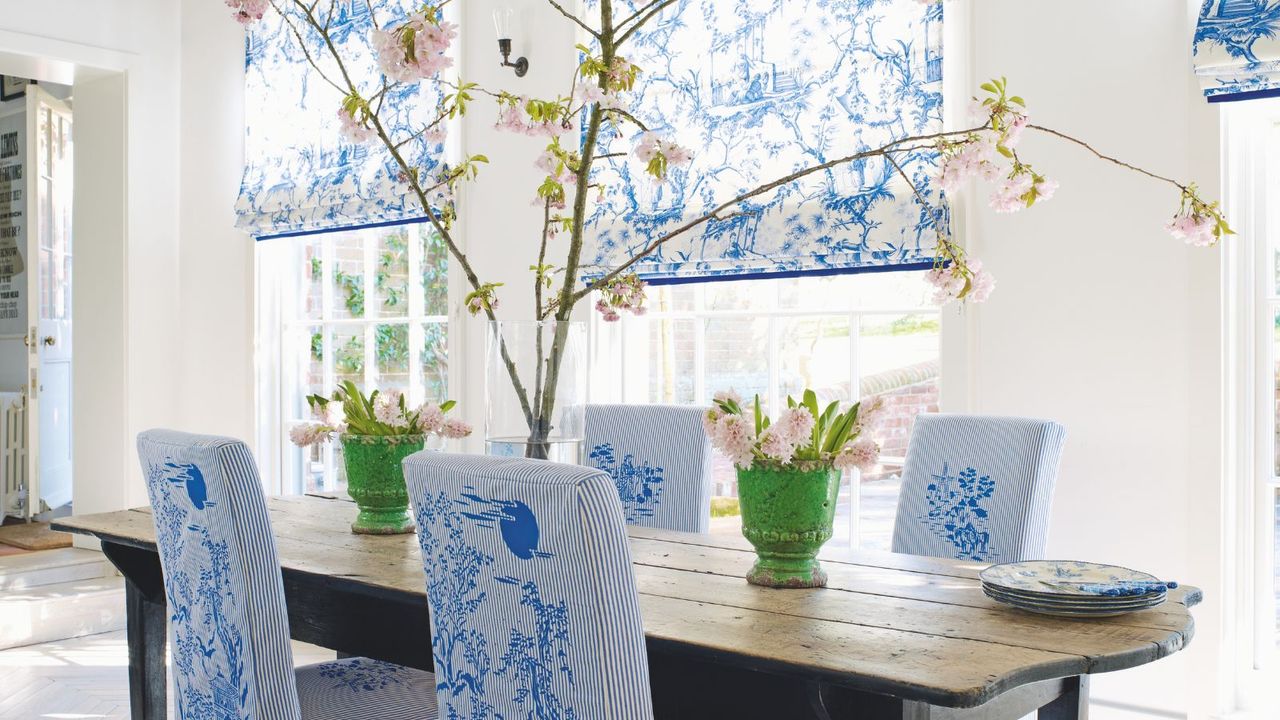 A spring dining room with a large wooden dining table in the centre, with pink flowers in green vases, and a large branch in the center of the table. Surrounding the table are four upholstered chairs in a blue and white Toile de Jouy pattern, and behind are large windows with matching patterned blinds. The walls in the dining room are white.