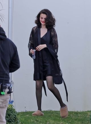 A photo of Selena Gomez wearing a black slip dress and a black lace robe with brown Ugg slippers on the set of her new music video.